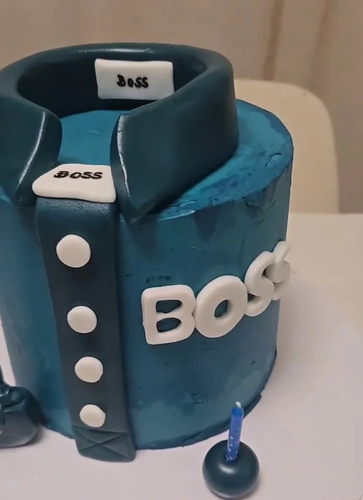 Gâteau d'anniversaire homme en forme de veste Hugo Boss : crème bleu pétrole, col tailleur bleu nuit, boutons blancs en relief, étiquette BOSS au col, lettres BOSS blanches sur le flanc, bougie bleue et petit sac en pâte à sucre