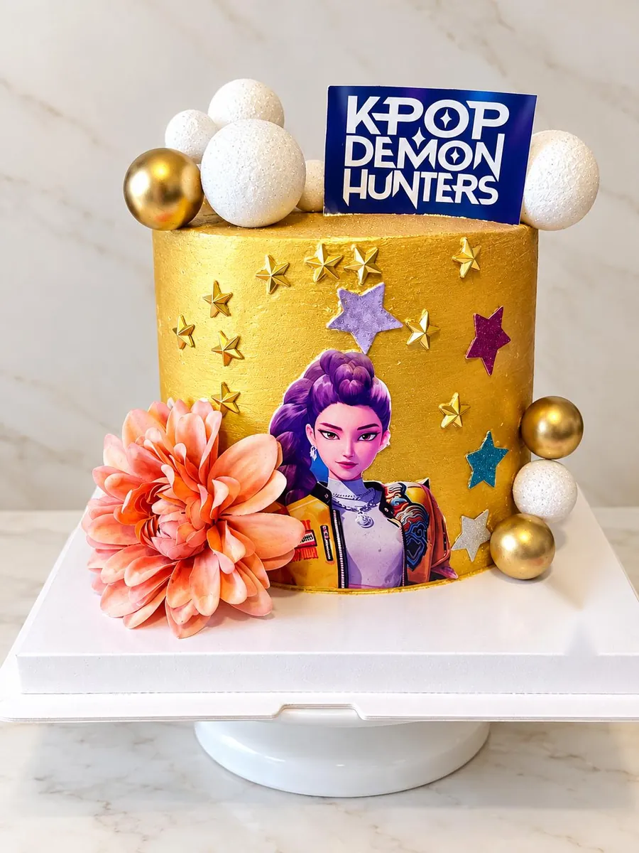 Gâteau KPop Demon Hunters : crème dorée texturée, étoiles dorées et pailletées, héroïne aux cheveux violets imprimée, topper KPop Demon Hunters, sphères blanches et dorées et grande fleur corail