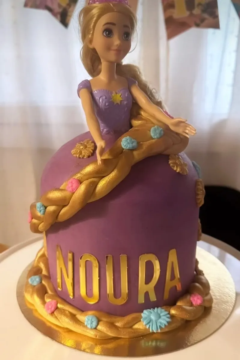 Gâteau poupée façon Raiponce : la robe violette est le gâteau, longue tresse dorée semée de fleurs, prénom Noura en lettres dorées